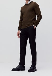 Pull en tricot brun foncé, pantalon noir ajusté et bottes en cuir noir poli. Détail des poignets de chemise blanche visibles aux poignets.