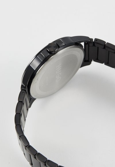 Montre-bracelet en métal noir avec un boîtier rond lisse, des accents polis et un bracelet à maillons. Elle dispose d'une couronne sur le côté droit et d'un dos en argent.