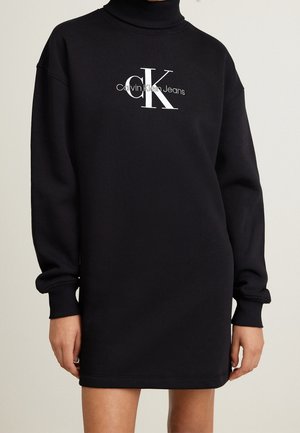 Czarna sukienka z długim rękawem i golfem, z białym logo "Calvin Klein Jeans" na środku klatki piersiowej, noszona przez osobę stojącą z rozluźnionymi ramionami.