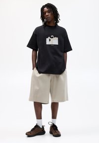 Schwarzes oversized T-Shirt mit einem grafischen Patch, kombiniert mit beigen Shorts und braunen Sportschuhen. Kurze Ärmel und lockere Passform.