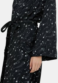 Schwarzer Bademantel mit weißem Sternmuster. Verfügt über lange Ärmel, einen gebundenen Taillengürtel und eine Tasche auf der linken Seite. Strukturierter Stoff.