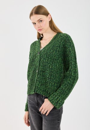 Young woman wearing a green textured knit cardigan with black buttons and dark gray jeans, standing against a plain white background.
