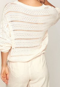 Pull en maille crème, présentant des rayures ajourées. Texture douce avec une coupe ample et des manches retroussées, associé à un pantalon en lin de couleur claire.