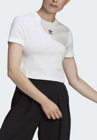 Balta trumpa medvilnės marškinėliai su trumpomis rankovėmis, ant krūtinės yra mažas juodas Adidas logotipas, derinami su juodomis plisuotomis kelnėmis.