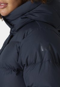 Chaqueta acolchada azul marino con un cuello alto, con una textura suave y un sutil logo brillante en la manga. La costura es visible a lo largo de las costuras.