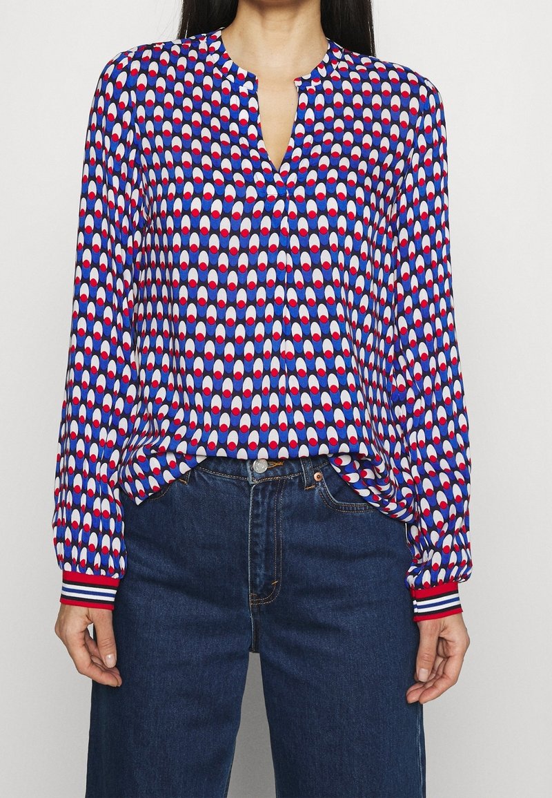 Personne portant un chemisier à manches longues avec un motif ovale bleu, blanc et rouge et des poignets rayés, rentré dans un jean taille haute bleu foncé.
