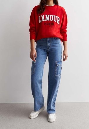 Kvinde iført en rød sweatshirt med teksten "L'AMOUR PARIS", blå cargobukser og hvide sneakers, stående mod en ensfarvet baggrund.