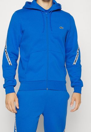Mann trägt eine leuchtend blaue Kapuzenjacke mit Reißverschluss und passende Hosen mit weißen Lacoste-Logo-Streifen an Ärmeln und Hosenbeinen.