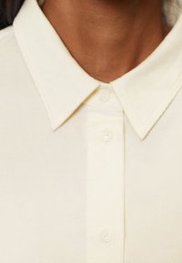 Camisa de botón color crema con un cuello de punta, que presenta una textura suave y cuatro botones a lo largo de la tapeta.