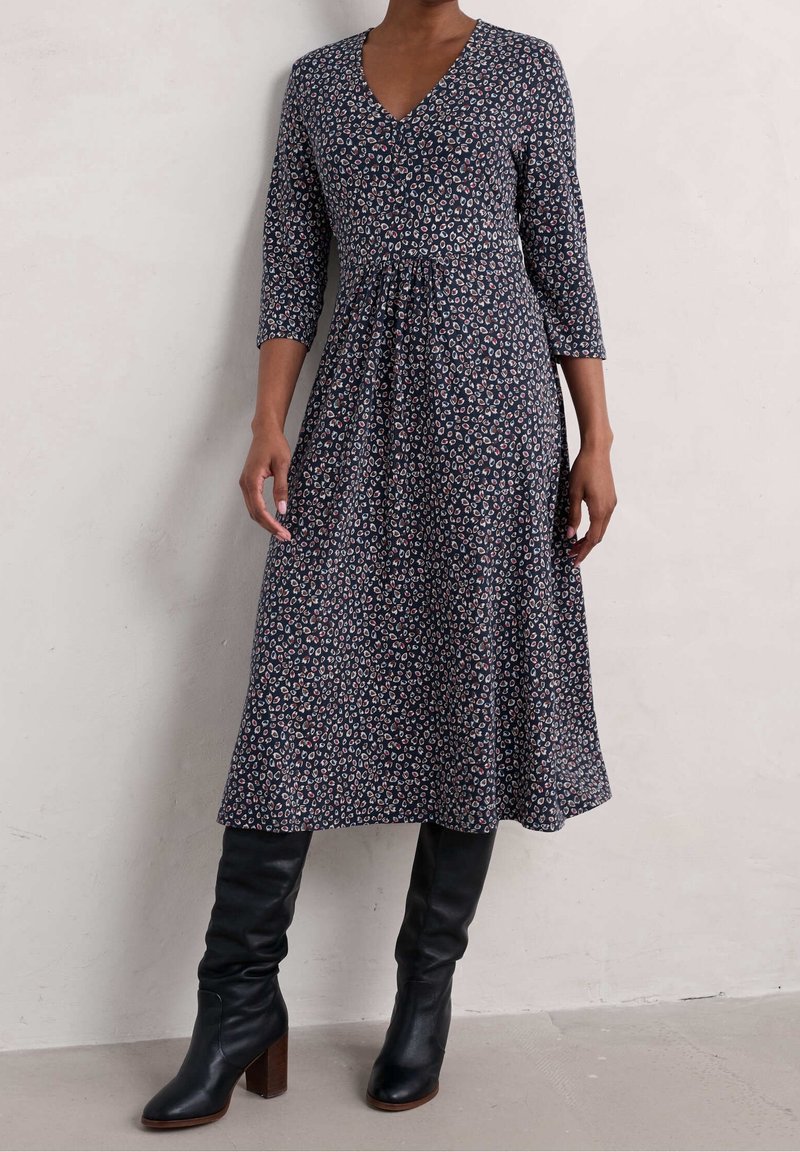 Femme portant une robe midi noire à manches longues à motifs, avec un col en V et des bottes noires hautes à talons, debout contre un mur uni.