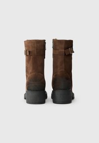 Brązowe zamszowe botki o długości do łydki, z dwoma bocznymi zamkami, z akcentami klamrowymi i teksturowaną czarną gumową podeszwą.