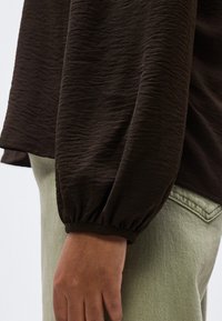 Nærbillede af en arm iført en mørkebrun tekstureret langærmet trøje med rynket manchet, sammen med lyse olivengrønne bukser med synlig lomme søm.