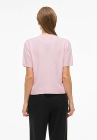 Džemper svijetlo ružičaste boje s kratkim rukavima, mekanog je teksture i ima rebrasti rub, okrugli izrez i opušten kroj straga.