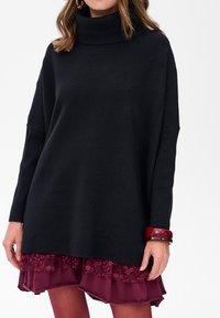 Zwarte coltrui met geribbelde details, gecombineerd met een gelaagde bordeauxrode rok met kanten trim. Geaccessoriseerd met een rode armband.