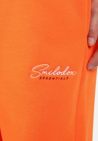 Pomarańczowe spodnie sportowe wykonane z gładkiego materiału, z białym haftowanym napisem "Smilodox ESSENTIALS" blisko szwu bocznego.