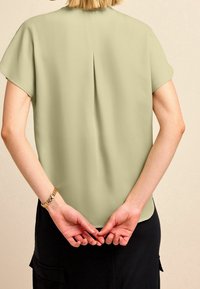 Helles grünes kurzärmeliges Blusenshirt mit einem Rückenfalten, glatter Textur, abgerundetem Saum und lockerer Passform, getragen mit dunklen Hosen.