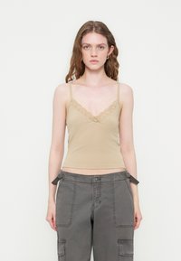 Jeune femme aux cheveux ondulés portant un caraco beige orné de dentelle et un pantalon cargo gris, debout devant un fond blanc uni.