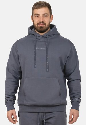 Mann trägt einen passenden dunkelgrauen Hoodie und Jogginghose mit Fronttasche und Kordeln, steht vor einem einfarbigen hellen Hintergrund.