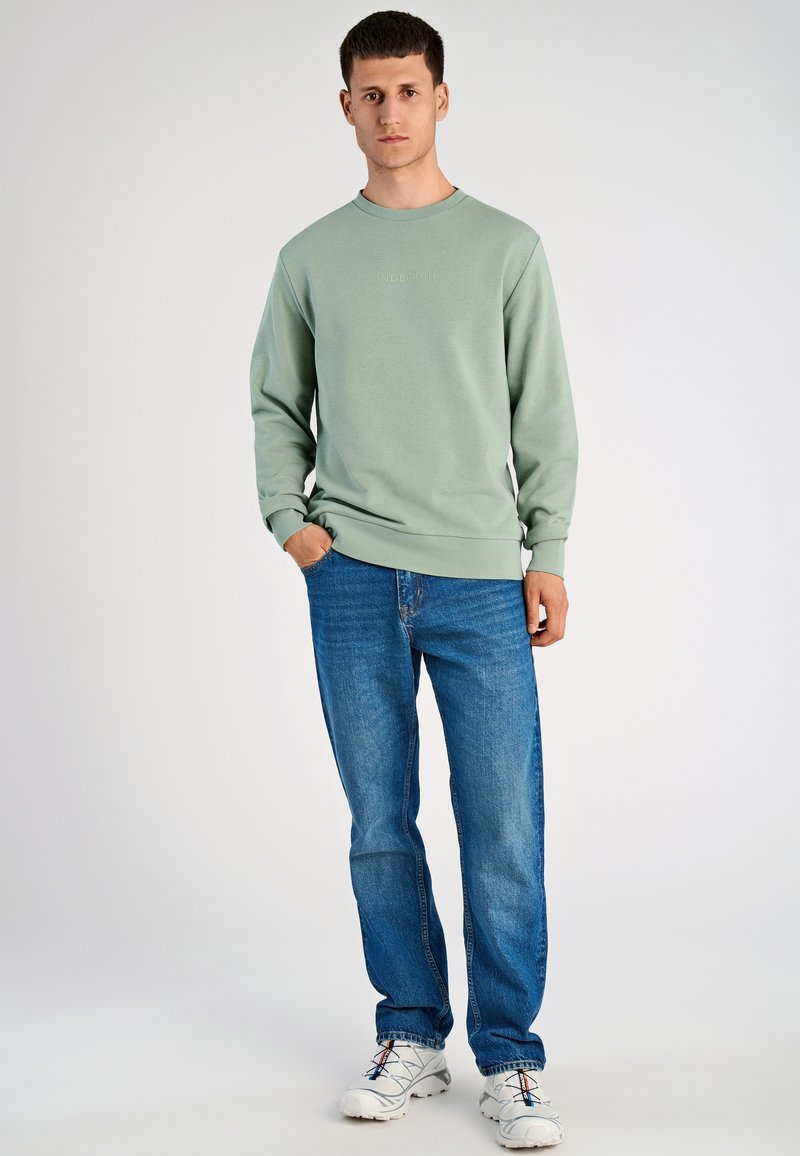 Felpa verde chiaro con collo rotondo e maniche svasate, abbinata a jeans blu e sneakers bianche. Dettaglio logo semplice sul davanti.