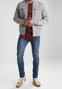 Gray bomber jacket with two chest pockets, maroon t-shirt, blue denim jeans, and beige sneakers with brown accents and white sole.