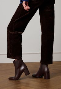 Braune Leder-Stiefeletten mit Blockabsatz und spitzer Zehenpartie; seitlicher Reißverschluss; getragen zu dunkelbraunen Cordhosen.