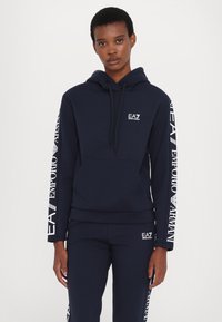 Sudadera de color azul marino con un bolsillo canguro, cordón y logo de EA7. Presenta una marca blanca a lo largo de las mangas, fabricada con un material suave.