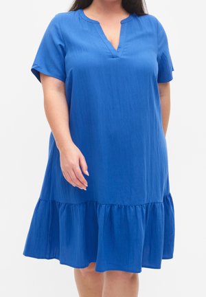 Kvinde iført en løstsiddende, kortærmet blå kjole med V-hals og en rynket, flæsekant over knæene.