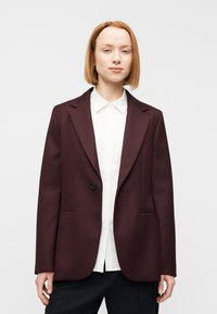 Μπορντό σακάκι με δομημένο σχεδιασμό, ένα κουμπί, δύο μπροστινές τσέπες και υφή με ανάγλυφο φινίρισμα, φορεμένο πάνω από λευκό πουκάμισο με γιακά.