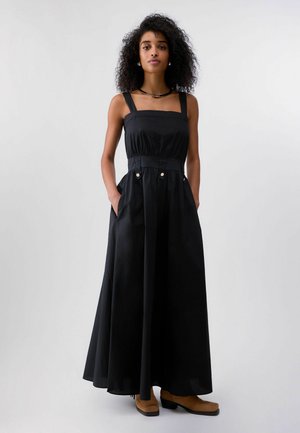 Lokkis juustega naine seisab näoga ettepoole, kandes varrukateta musta maksikleit nööpidega ja beeže pahkluukombeid.