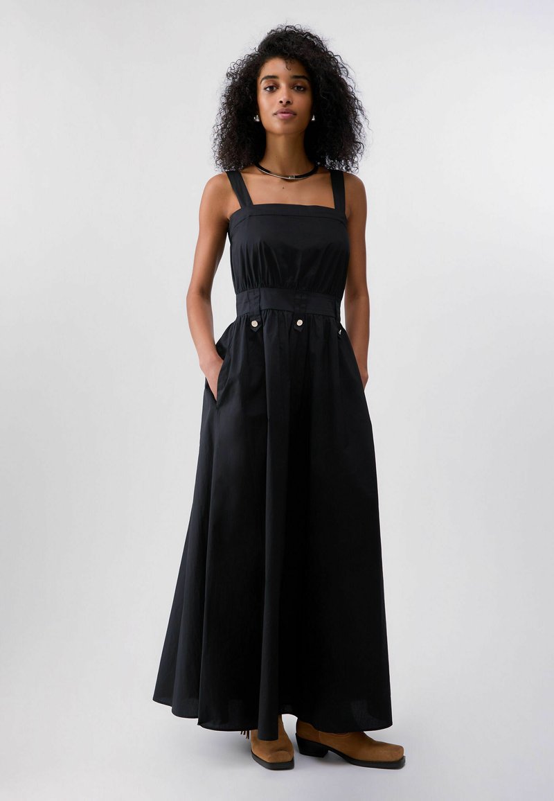 Femme aux cheveux bouclés, debout de face, portant une robe maxi noire sans manches avec des détails à boutons et des bottines camel.