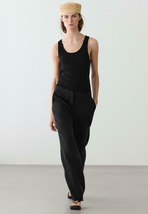 Femme portant un bandeau tissé beige, un haut noir sans manches, un pantalon noir ample et des sandales noires, marchant devant un fond blanc uni.