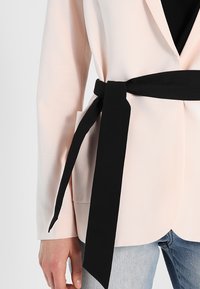 Jasnoróżowa marynarka z czarnym materiałowym paskiem, o dopasowanym kroju, gładkiej fakturze i klasycznym kołnierzu z klapami.