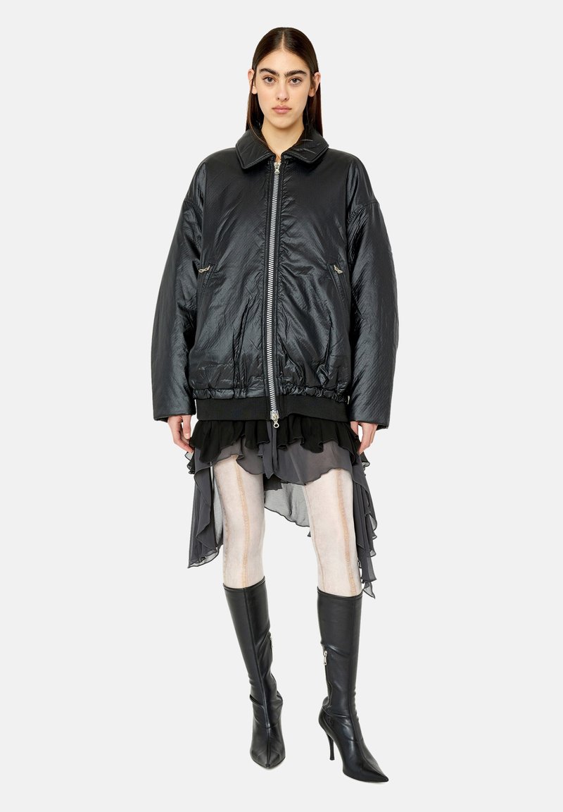 Frau steht in einer schwarzen oversized Zip-up-Jacke, mit einem geschichteten, gerafften Rock, hellen Strumpfhosen und schwarzen knielangen Stiefeln mit Absatz vor einfarbigem Hintergrund.