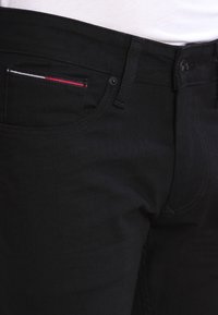 Jeans de mezclilla negra con una textura suave, corte ajustado y un pequeño detalle a rayas en rojo, blanco y azul en el bolsillo.