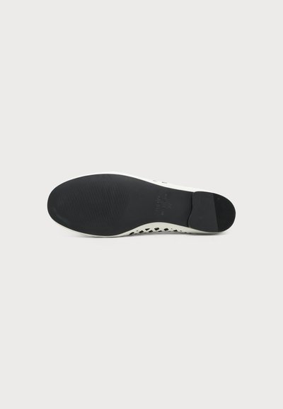 Semelle noire d'une chaussure plate blanche vue de dessous avec des zones texturées aux orteils et un talon solide sur un fond neutre.