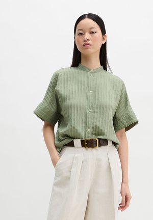 Kobieta w jasnozielonej krótkorękawowej koszuli zapinanej na guziki, włożonej do beżowych spodni z wysokim stanem i brązowym paskiem, stojąca na białym tle.