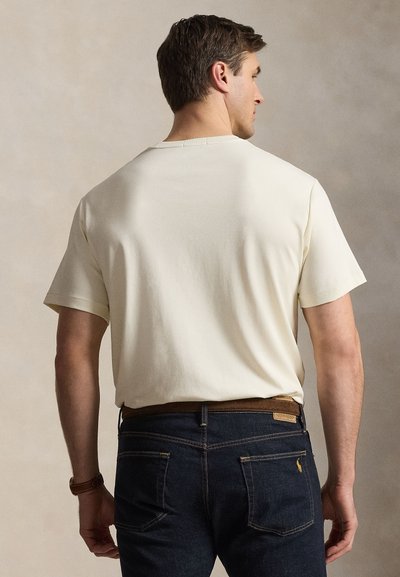 Hátulról látható férfi, egyszínű törtfehér rövid ujjú pólót visel, amelyet sötétkék farmerba tűrt barna övvel, semleges háttér előtt.