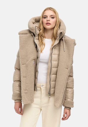 Femme portant des vestes matelassées et en polaire beige superposées sur un haut blanc et un pantalon crème clair, debout devant un fond uni.