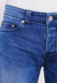 Jeans de mezclilla azul con corte regular, que presentan un diseño clásico de cinco bolsillos, herrajes metálicos y un ligero desvanecimiento en la tela.