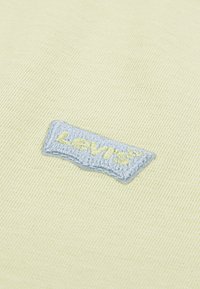 Κίτρινο ύφασμα με υφή ύφανσης διαθέτει ένα ανοιχτό μπλε κεντημένο έμβλημα με το λογότυπο της Levi's. Ο σχεδιασμός είναι ορθογώνιος με στρογγυλεμένες άκρες.