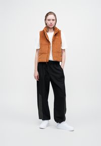 Oranger Cord-Vest über einem weißen T-Shirt, kombiniert mit schwarzen Hosen mit olivgrünen Akzenten und weißen Sneakers.