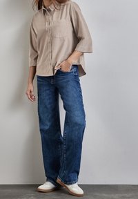 Camicia beige con bottoni e taschino sul petto, abbinata a jeans larghi blu e sneakers bianche, su sfondo neutro.