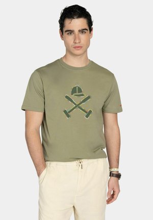 Joven con camiseta verde oliva con gráfico de bates de béisbol cruzados y gorra, combinada con pantalones con cordón de color crema.