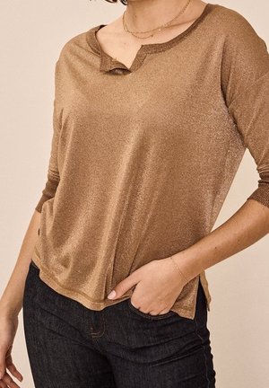 Třpytivý bronzový top s rukávy tři čtvrtě dlouhými a malým výstřihem s vyříznutím, nošený s tmavě modrými džíny a jemnými zlatými šperky na zápěstí a krku.