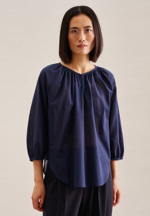 Blusa leggera di colore blu navy con scollo rotondo, dettagli arricciati sulle spalle e maniche lunghe leggermente arricciate.