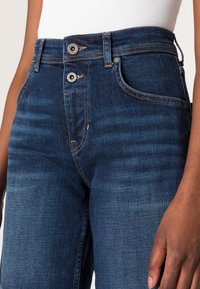 Jeansy z wysokim stanem w kolorze niebieskim, z dwoma przednimi guzikami, delikatnym przecieraniem, subtelnymi szwami oraz pięcioma kieszeniami. Gładka faktura denimu.