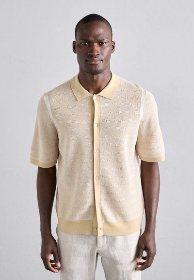 Kortærmet strikket polo t-shirt i lys beige med gule accenter, som har et struktureret mønster, knapper og en blød krave.