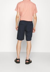 Man i korallrandig kortärmad skjorta, marinblå shorts och svarta sandaler, står vänd bort från kameran på en enfärgad vit bakgrund.