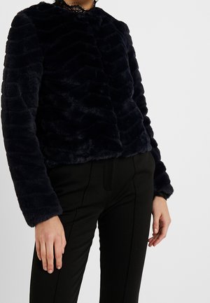 Person wearing a black faux fur long-sleeve jacket and black tailored pants, standing against a plain light background.