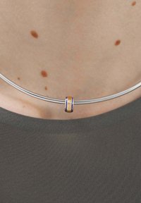 Collar plateado con un acento dorado y rayas azules, descansando sobre un fondo de tela gris. El diseño es liso y minimalista.